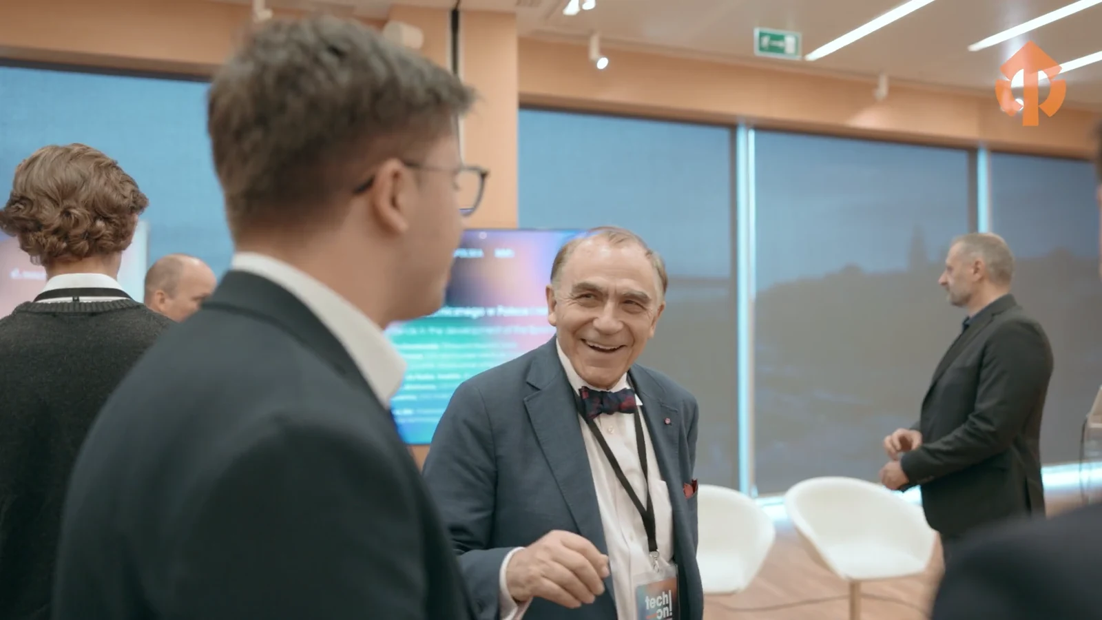 Uczestnicy konferencji rozmawiają w kuluarach, w tle ekran z prezentacją