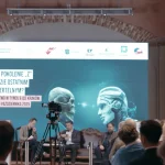Widownia konferencji, na scenie ekran z prezentacja, przed publicznoscia kamera na statywie